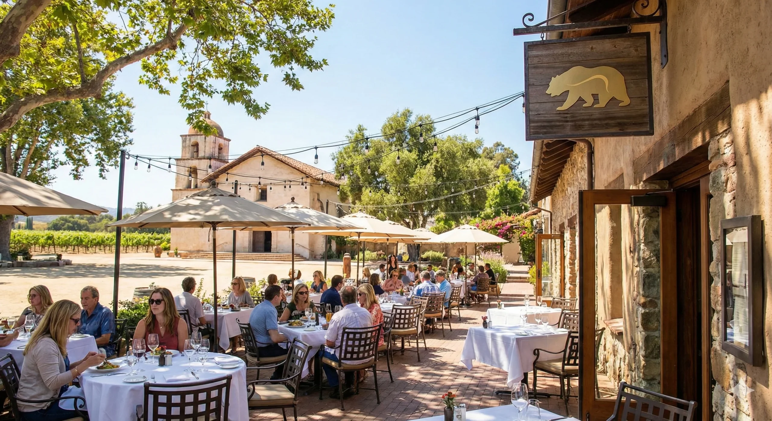 A bustling outdoor dining patio at el dorado kitchen, featuring white-clothed tables, umbrellas, and people enjoying meals under a golden bear sign, with a mission church and vineyard in the background.